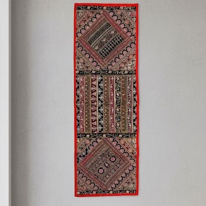 May include: A long, rectangular textile wall hanging with a red border. The central design features intricate patterns in black, red, and beige. The wall hanging is displayed on a light grey wall, adding a touch of bohemian style to the room.