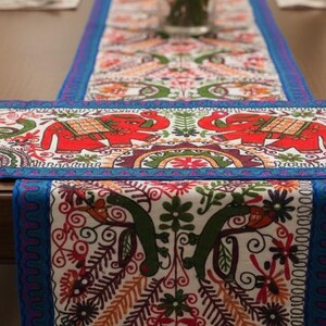 Könnte beinhalten: Ein rechteckiger, bestickter Tischläufer mit blauem Rand und aufwendigen Mustern. Das Design zeigt rote Elefanten, florale Motive und ein zentrales Muster. Der Läufer liegt auf einem dunklen Holztisch und erzeugt einen Kontrast von Farben und Texturen.