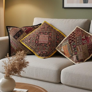 Puede incluir: Tres cojines decorativos con intrincados y coloridos diseños descansan sobre un sofá gris claro. Los cojines presentan una mezcla de tonos burdeos, dorados y negros. Una mesa de centro redonda de madera y una alfombra beige completan la escena.