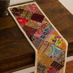 Può includere: Una colorata tovaglia patchwork con bordo color crema. La tovaglia presenta una varietà di motivi in tessuto nei toni del rosso, oro, blu e rosa. È appoggiata su un tavolo di legno scuro.