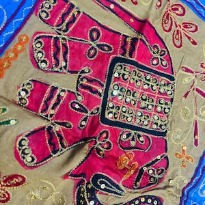 Könnte beinhalten: Dekoratives Textil mit einem gestickten Elefantenmuster. Der Elefant ist hauptsächlich rosa mit schwarzen und goldenen Details und mit Pailletten verziert. Der Hintergrund ist beige, mit einem blauen Rand. Es handelt sich wahrscheinlich um einen dekorativen Kissenbezug oder einen Wandbehang.