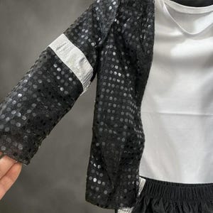 Puede incluir: Una chaqueta negra de lentejuelas con una banda plateada brillante en la manga, sobre una camiseta blanca lisa. La chaqueta est&aacute; confeccionada con una tela negra semitransparente cubierta densamente por lentejuelas redondas y reflectantes.