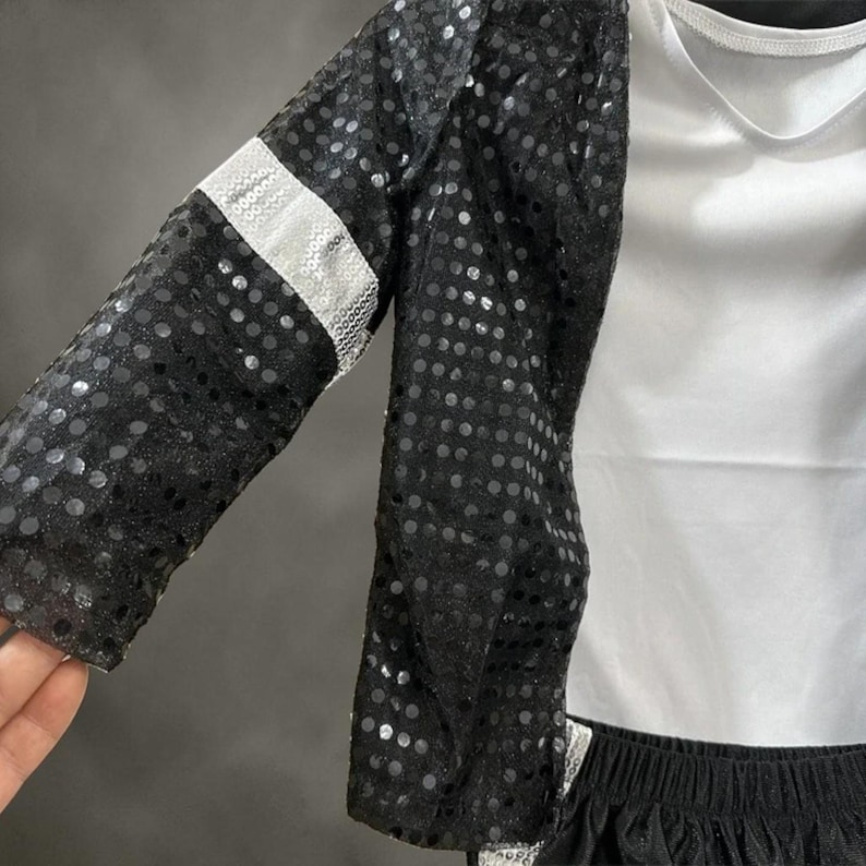 Puede incluir: Una chaqueta negra de lentejuelas con una banda plateada brillante en la manga, sobre una camiseta blanca lisa. La chaqueta est&aacute; confeccionada con una tela negra semitransparente cubierta densamente por lentejuelas redondas y reflectantes.