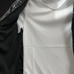 Puede incluir: Una camiseta blanca lisa debajo de una chaqueta negra abierta. La chaqueta presenta una superficie exterior cubierta densamente con peque&ntilde;as lentejuelas negras reflectantes y un forro de tela negra s&oacute;lida en el interior, creando un contraste.