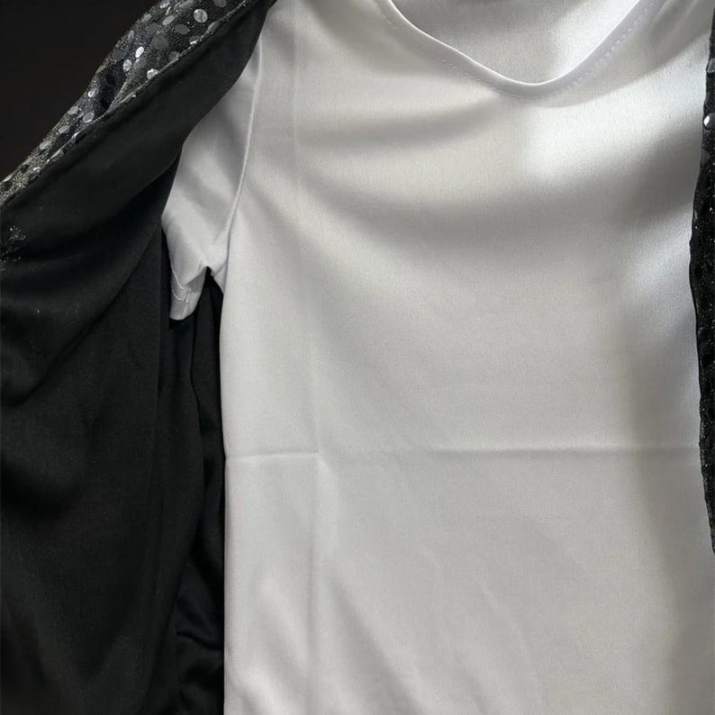 Puede incluir: Una camiseta blanca lisa debajo de una chaqueta negra abierta. La chaqueta presenta una superficie exterior cubierta densamente con peque&ntilde;as lentejuelas negras reflectantes y un forro de tela negra s&oacute;lida en el interior, creando un contraste.