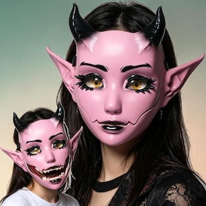 Puede incluir: Una máscara de diablo rosa con cuernos negros, orejas puntiagudas y grandes ojos amarillos con largas pestañas negras. La máscara tiene un acabado liso, labios negros y una mandíbula marcada. Se muestra una versión más pequeña del diseño.