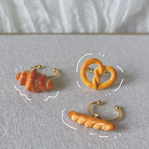 Peut inclure: Trois bagues réglables avec des breloques miniatures en forme d'aliments : un croissant, un bretzel et une baguette. Les breloques sont détaillées avec des couleurs et des textures réalistes, fixées sur des bagues réglables dorées.