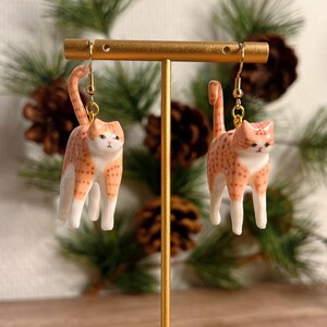Op de afbeelding: Een paar katvormige oorbellen. Elke oorbel heeft een kleine, oranje en witte kat met een gevlekt patroon. De katten staan rechtop met hun staarten omhoog. De oorbellen hangen aan goudkleurige haken.