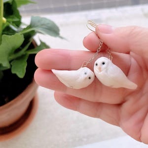 Op de afbeelding: Een paar witte uilen oorbellen. Elke oorbel heeft een kleine, gebeeldhouwde uil met zwarte ogen en een delicate ketting. De oorbellen worden omhoog gehouden door een goudkleurige haak. De achtergrond bevat een groene plant in een bruine pot.