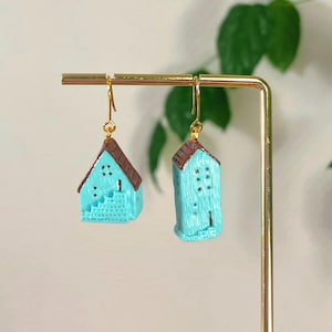 Peut inclure: Boucles d'oreilles en forme de maison turquoise avec des toits bruns et des crochets dorés. Les boucles d'oreilles miniatures présentent de petites fenêtres et des détails architecturaux. Elles sont exposées sur un présentoir doré. Hauteur : environ 2 cm.