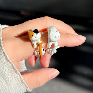Op de afbeelding: Twee katvormige ringen. Eén ring heeft een witte kat met een lachend gezicht. De andere ring heeft een lapjeskatontwerp met oranje, zwarte en witte kleuren. Beide ringen zijn gemaakt van een glanzend materiaal.