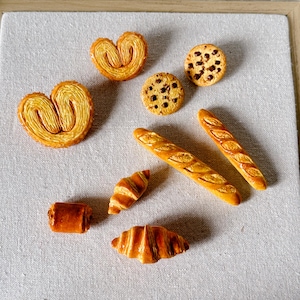 Peut inclure: Assortiment de miniatures de boulangerie, comprenant croissants, baguettes, cookies et palmiers, disposées sur une surface claire. Les pâtisseries sont brun doré avec des textures détaillées, suggérant un thème de boulangerie.
