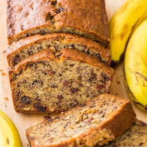 Può includere: Un pane alla banana con noci e frutta secca scura visibili, affettato in diversi pezzi. Il pane ha una crosta dorata e un interno strutturato. Tre banane gialle intere sono posizionate accanto al pane affettato.