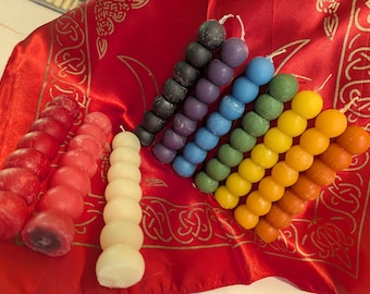 May include: A collection of colorful, beaded candles in shades of red, white, black, purple, blue, green, yellow, and orange. The candles are arranged on a red cloth with a gold Celtic knot design.