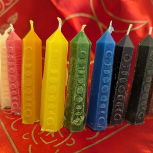 Puede incluir: Una colección de ocho velas hexagonales de colores. Las velas están dispuestas en fila, con colores que van desde el blanco, rosa, amarillo, verde, azul, negro y marrón. Cada vela tiene una mecha y un patrón de fases lunares.