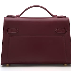 May include: A structured burgundy handbag with a single top handle, a flap closure, and two side straps. The bag features a smooth finish, a rectangular silhouette, and small gold-toned metal feet on the bottom base for stability.