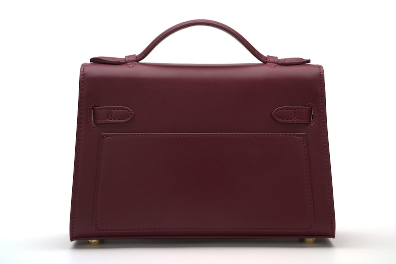 May include: A structured burgundy handbag with a single top handle, a flap closure, and two side straps. The bag features a smooth finish, a rectangular silhouette, and small gold-toned metal feet on the bottom base for stability.