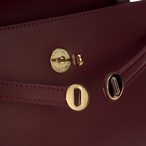 May include: A deep burgundy handbag featuring gold-toned hardware, including two horizontal straps with circular metallic clasps and a central turn-lock closure. The circular lock is engraved with the text CHOOSE CONSCIOUSLY.