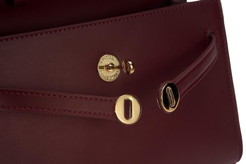 May include: A deep burgundy handbag featuring gold-toned hardware, including two horizontal straps with circular metallic clasps and a central turn-lock closure. The circular lock is engraved with the text CHOOSE CONSCIOUSLY.