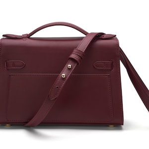 May include: A deep burgundy leather handbag featuring a structured rectangular body, a top handle, and an adjustable shoulder strap with gold-toned metal hardware. The bag includes a flap closure with side strap details and a smooth finish.