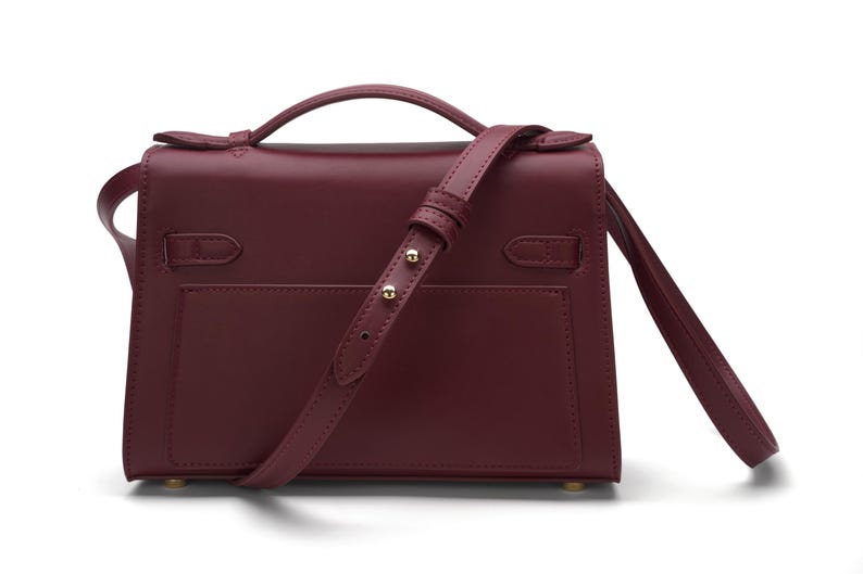 May include: A deep burgundy leather handbag featuring a structured rectangular body, a top handle, and an adjustable shoulder strap with gold-toned metal hardware. The bag includes a flap closure with side strap details and a smooth finish.