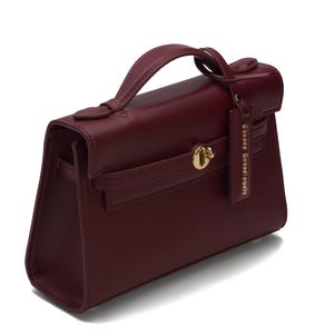 May include: A deep burgundy handbag with a structured silhouette, a single top handle, and a gold-toned turn-lock closure. A matching rectangular leather tag hangs from the handle, featuring the embossed text "GEORGE GINA & LUCY".