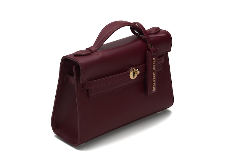 May include: A deep burgundy handbag with a structured silhouette, a single top handle, and a gold-toned turn-lock closure. A matching rectangular leather tag hangs from the handle, featuring the embossed text "GEORGE GINA & LUCY".