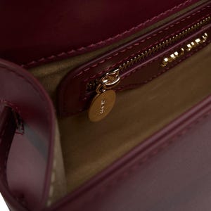 May include: A deep burgundy leather handbag with a smooth finish, featuring a beige interior lining, a gold-toned zipper closure with a circular gold metal pull tab, and a gold-toned brand logo plate mounted above the interior pocket.