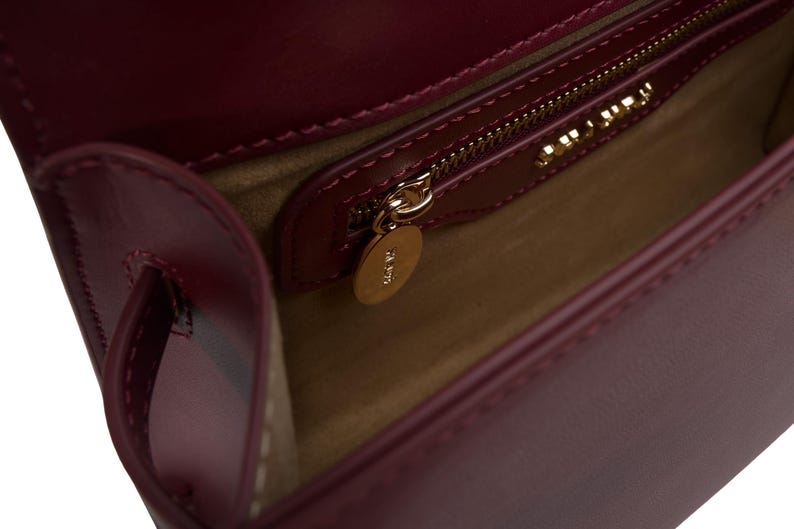 May include: A deep burgundy leather handbag with a smooth finish, featuring a beige interior lining, a gold-toned zipper closure with a circular gold metal pull tab, and a gold-toned brand logo plate mounted above the interior pocket.