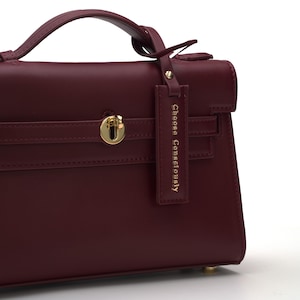 May include: A deep burgundy handbag featuring a top handle, a gold-toned metal turn-lock closure, and a rectangular hanging tag with the text "Choose Consciously" embossed in gold. The bag has a structured design with clean, smooth lines.