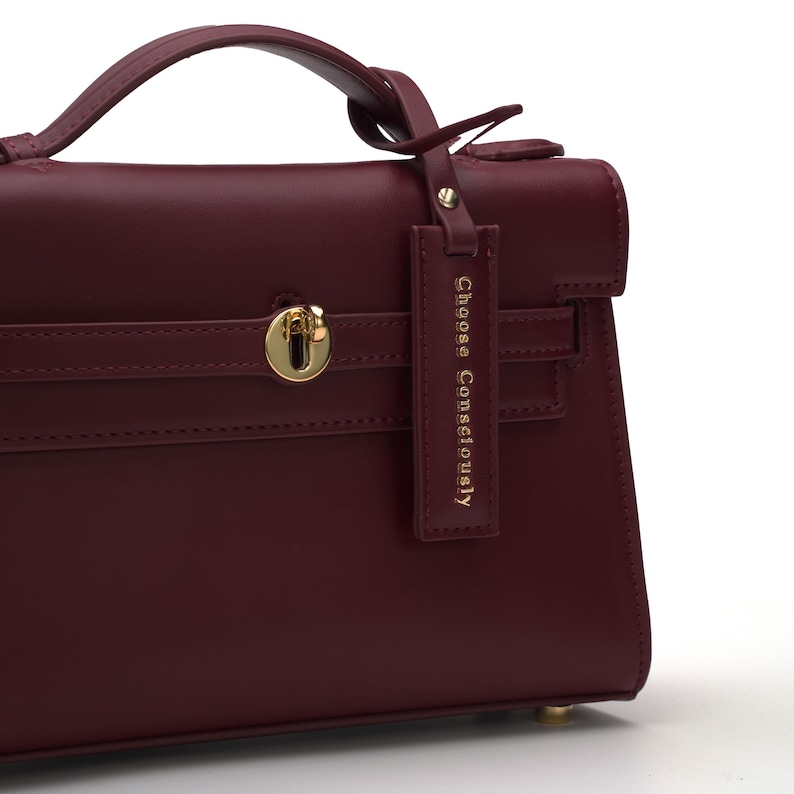 May include: A deep burgundy handbag featuring a top handle, a gold-toned metal turn-lock closure, and a rectangular hanging tag with the text "Choose Consciously" embossed in gold. The bag has a structured design with clean, smooth lines.