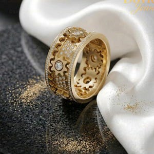 May include: A gold-colored ring with a gear design, encrusted with small, clear gemstones. The ring features a series of interlocking gears around its band, with a central gear design. The ring is displayed on a reflective surface.