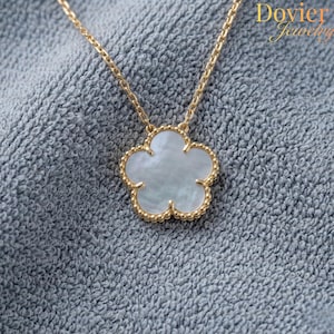 Op de afbeelding: Een goudkleurige ketting met een bloemvormige hanger. De hanger heeft een witte, parelmoerachtige kern en een gouden, kralenrand. De ketting is ook goudkleurig. De achtergrond is een grijze, getextureerde stof.