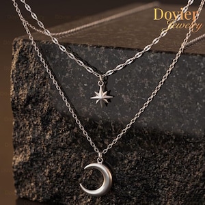 Pode incluir: Um colar prateado em camadas com um pingente de estrela e um pingente de lua crescente. O colar é exibido em uma superfície escura e texturizada. A camada superior tem uma corrente com um design em forma de diamante.