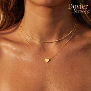 Könnte beinhalten: Eine goldfarbene Halskette mit zwei Strängen. Der obere Strang hat ein Perlendesign, und der untere Strang hat einen kleinen Herzanhänger. Der Markenname "Dovier Jewelry" ist oben rechts sichtbar.