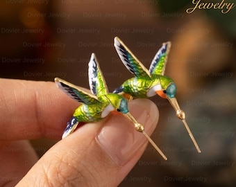 May include: A pair of hummingbird earrings. Each earring features a green and gold body with blue, black, and white wings. The earrings have long, thin gold beaks and are held by a hand.
