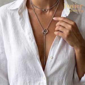 May include: A silver necklace with two layers. The top layer is a choker with a single ring pendant. The second layer has a lariat design with two interlocking rings and a vertical bar pendant. The necklace is worn over a white button-down shirt.