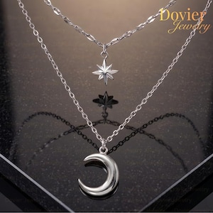 May include: A silver layered necklace featuring a crescent moon pendant and a starburst pendant. The necklace has two chains, one with a delicate link design and the other with a classic chain. The jewelry is displayed on a dark surface.