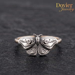 Peut inclure: Une bague argentée présentant un motif de papillon de nuit détaillé en pièce centrale. Le papillon possède des ailes texturées aux motifs complexes et un corps lisse. L'anneau est fin et métallique, positionné horizontalement sur un fond sombre.