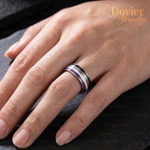 May include: A metallic band ring featuring a horizontal inlay with stripes of purple, white, gray, and black. The ring has a polished finish and is worn on a finger. The text Dovier Jewelry is visible in the upper right corner of the image.