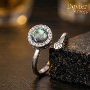 May include: A silver ring featuring a round, iridescent abalone shell center stone surrounded by a halo of small, clear gemstones. A smaller, clear gemstone is set on the open band. The ring is displayed on a dark, textured surface.