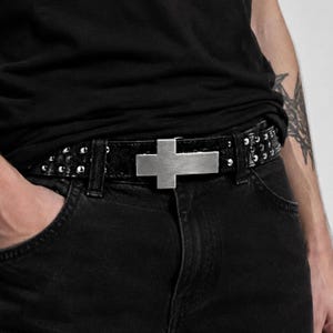 Peut inclure: Une ceinture noire texturée dotée d'une boucle en métal argenté brossé en forme de croix. La sangle de la ceinture est ornée de multiples petits clous métalliques argentés ronds disposés en rangées sur toute la longueur de la bande.