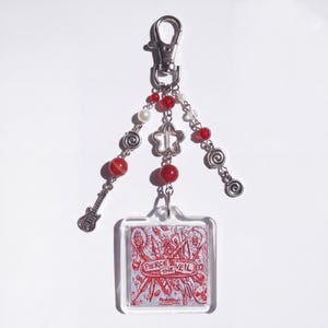 May include: A silver-toned metal keychain featuring a square acrylic charm with a red line-art illustration and the text "PIERCE THE VEIL". The charm hangs from a lobster clasp with three beaded chains containing red and white beads, silver spiral charms, and a small guitar charm.