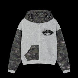 Pode incluir: Um casaco cinzento com fecho, mangas compridas e capuz com padrão de camuflagem verde escuro. A frente inclui um gráfico abstrato preto no peito e um bolso com padrão de camuflagem no lado inferior esquerdo.