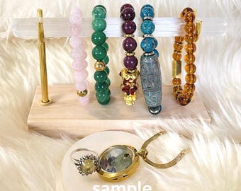 Könnte beinhalten: Eine Präsentation von Armbändern mit Perlen in verschiedenen Farben, darunter Rosa, Grün, Burgunderrot, Blau und Bernstein, auf einem Holz- und goldfarbenen Ständer. Ein Ring und ein goldfarbenes Medaillon mit einem Edelstein sind ebenfalls ausgestellt. Das Wort "sample" ist auf der Oberfläche aufgedruckt.