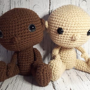 May include: Two crocheted dolls, one brown and one beige, sitting side by side. Both dolls have black eyes and are made with a textured stitch.