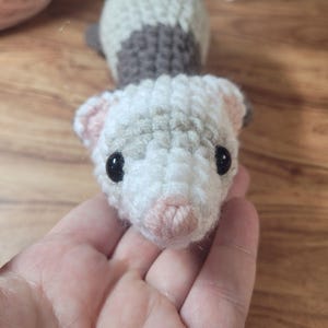 Puede incluir: Un hurón de ganchillo hecho a mano en blanco, gris y rosa. El hurón tiene ojos negros y se sostiene en una mano. El hurón está hecho de hilo y tiene un aspecto suave y texturizado. El fondo es una superficie de madera.