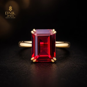 Puede incluir: Un anillo de oro con una gran gema rectangular de color rojo intenso. La gema está sujeta por cuatro garras de oro. La banda del anillo es de oro pulido. El fondo negro resalta el brillo del anillo.