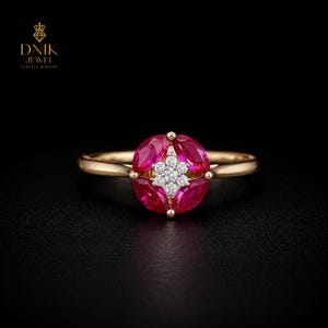 Op de afbeelding: Een gouden ring met een bloemmotief, met robijnen en een centrale cluster van diamanten. De ring heeft een gouden band en is geplaatst tegen een zwarte achtergrond. Het ontwerp van de ring is elegant en opvallend.