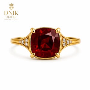 Op de afbeelding: Een gouden ring met een grote, kussengeslepen dieprode edelsteen vastgehouden door vier pootjes. De band is aan weerszijden van de zetting versierd met drie kleine, ronde, heldere steentjes. De tekst DNIK JEWEL PREMIUM JEWELRY staat erboven.